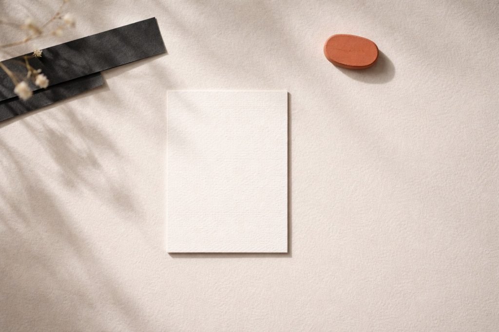 Carte blanche posée sur un fond texturé beige clair, ton sur ton, avec un galet corail et une bande noire en bordure